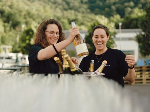 Zwei Frauen stehen vor Weingläsern und Sekflaschen. Eine Frau hält eine Flasche hoch. Beide lachen.