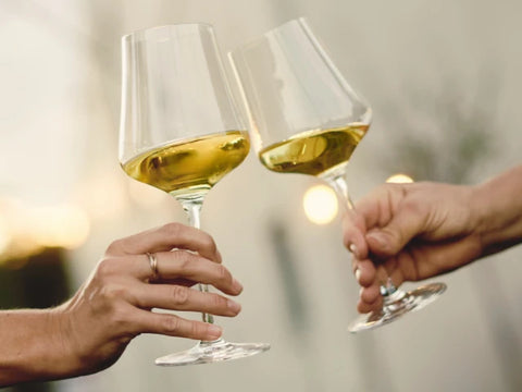 zwei Hände halten jeweils Ein Weinglas mit Weißwein. Sie stoßen an.